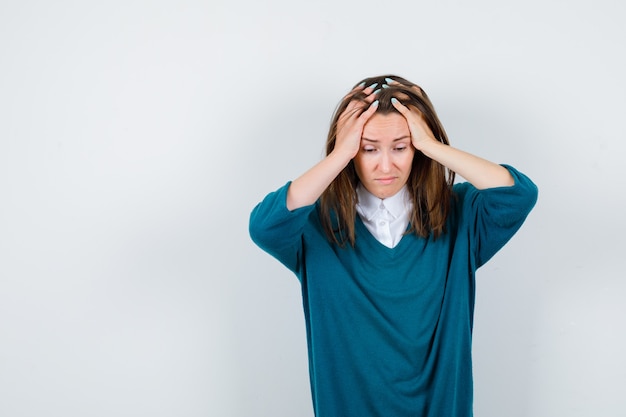 Young girl holding head with hands in v-neck sweater, shirt and looking dismal , front view.