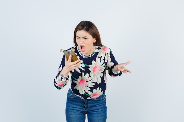 Young girl holding phone, being displeased with dumb question in floral blouse, jeans and looking resentful, front view.