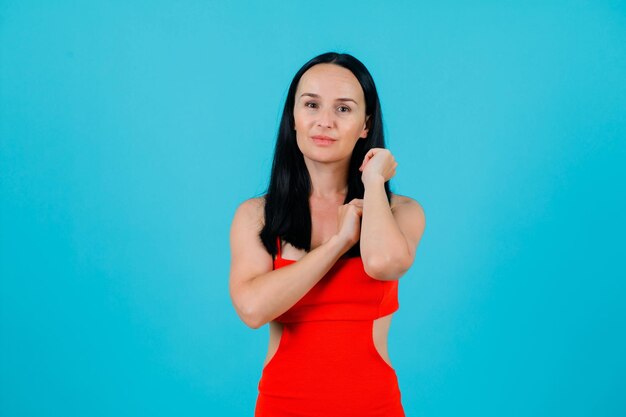 Young girl is looking at camera by holding hands near shoulder on blue background