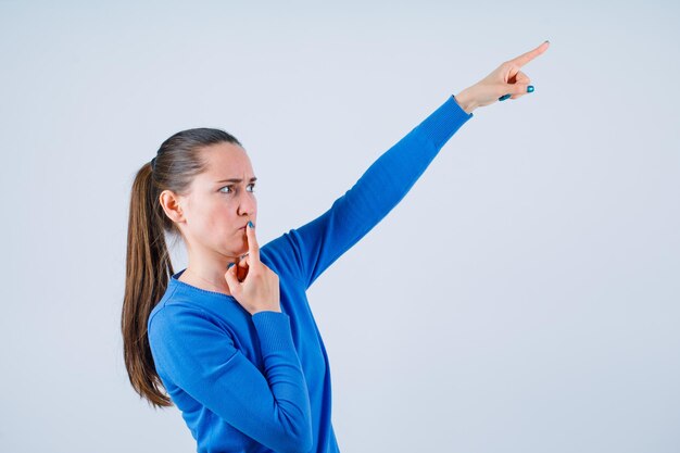 Young girl is pointing right up with forefinger and putting other forefinger on lips on white background