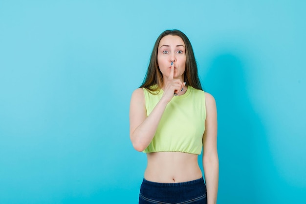 Young girl is showing silence gesture by holding forefinger on lips on blue background