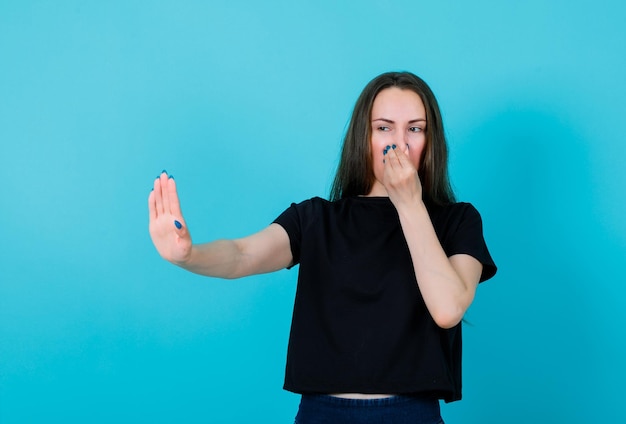 Young girl is showing stop gesture and holding nose with other hand on blue background