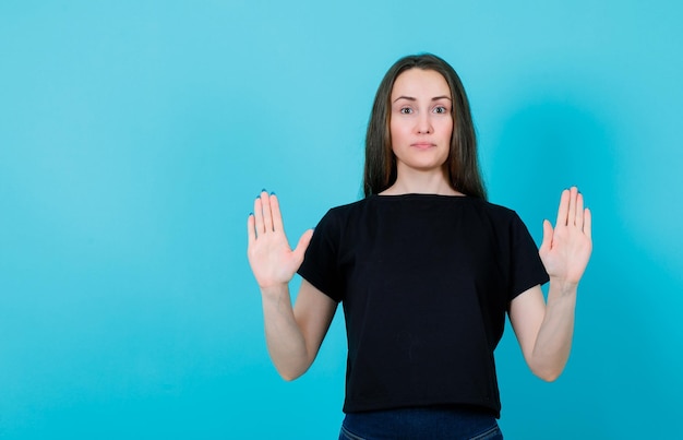 Young girl is showing stop gestures by raising up her hands on blue background