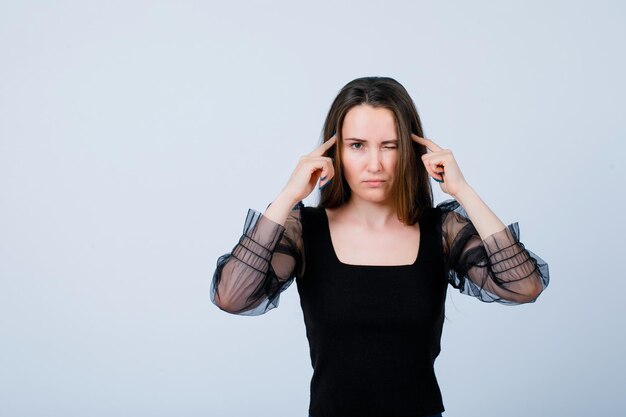 Young girl is winking by holding forefingers on temples on white background