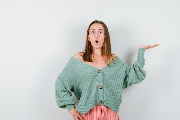 Young girl in knitwear, skirt holding hand on waist, spreading palm aside and looking surprised , front view.