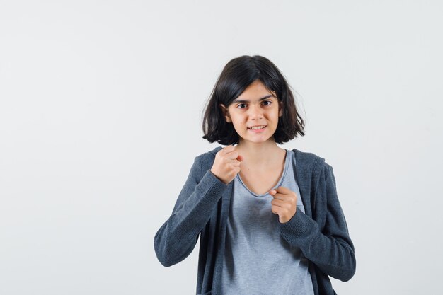 Young girl in light gray t-shirt and dark grey zip-front hoodie showing winner gesture and looking cute