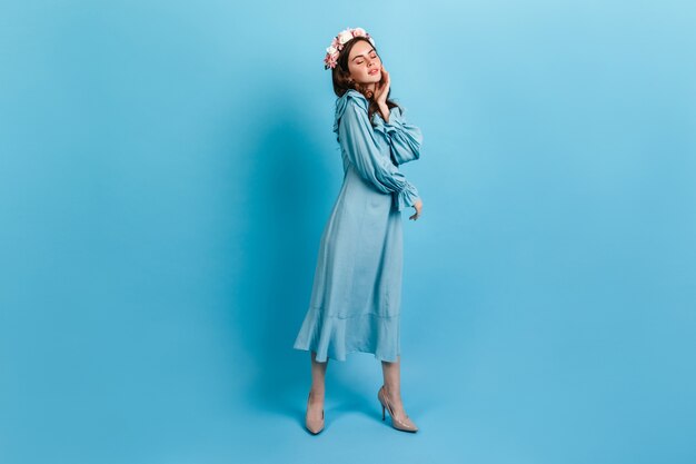 Young girl in long dress posing on blue wall. Model with roses in hair gently touches face.