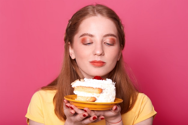 Free photo young girl, looks at plate with piece of birthday cake isolated on pink, wants to eat tasty dessert, wears yellow t shirt, has perfect hair style, poses.