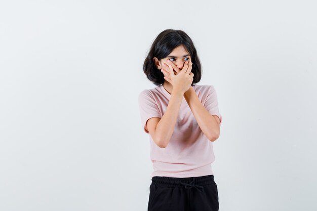 Young girl in pink t-shirt and black pants covering mouth with both hands and looking cute