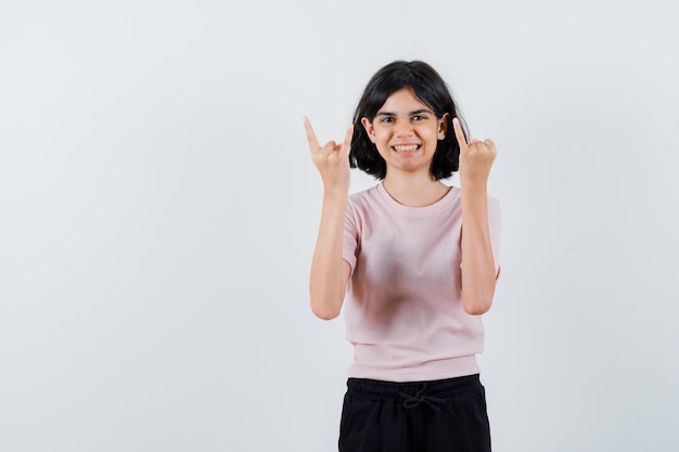 Young girl in pink t-shirt and black pants showing heavy metal gesture and pinky finger and looking happy