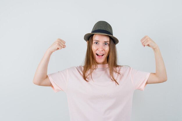 Young girl in pink t-shirt, hat showing winner gesture and looking happy , front view.