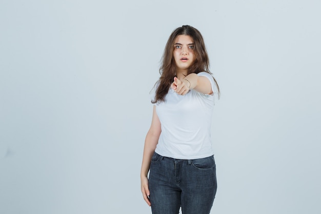 Young girl pointing at camera in t-shirt, jeans and looking surprised , front view.