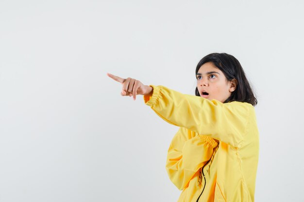 Young girl pointing left corner with index finger in yellow bomber jacket and looking surprised.