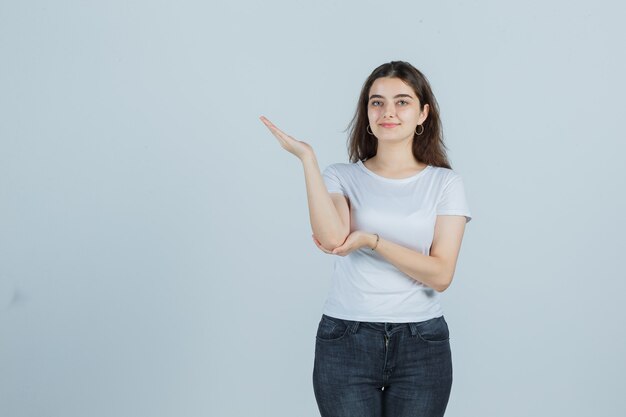 Young girl pretending to show something in t-shirt, jeans and looking joyful , front view.