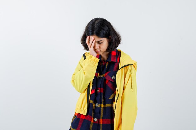 Young girl putting hand on face in checked shirt and yellow jacket and looking tired , front view.