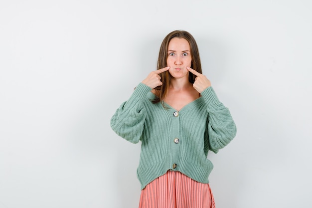 Young girl putting index finger on cheeks in knitwear, skirt and looking serious. front view.