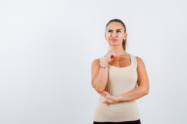 Young girl putting index finger under chin, looking upward in beige top, black pants and looking pensive , front view.