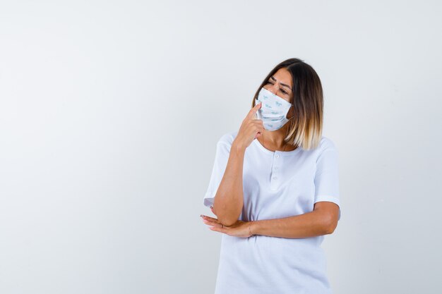 Young girl putting index finger on mouth, holding hand under elbow in white t-shirt and mask and looking pensive , front view.