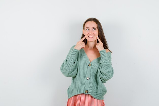 Young girl putting index fingers on cheek, looking away in knitwear, skirt and looking happy. front view.