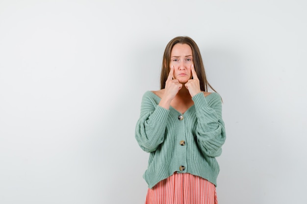 Young girl putting index fingers on cheeks in knitwear, skirt and looking morose , front view.