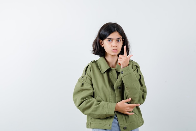 Young girl raising index finger in eureka gesture in grey sweater, khaki jacket, jean pant and looking cute. front view.