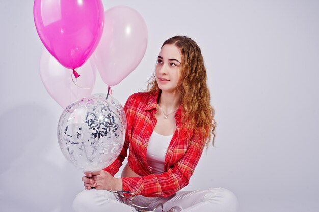 Young girl in red checked shirt and white pants with balloons against white background on studio