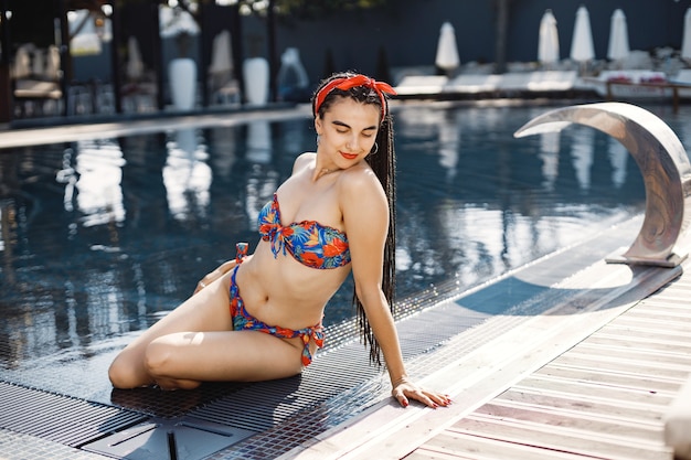 Young girl in a red headband sitting near swimming pool. Brunette girl wearing red and blue swimwear