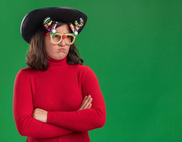 Free photo young girl in red sweater wearing funny glasses and black hat looking aside displeased with arms crossed blowing cheeks standing over green wall