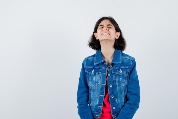 Young girl in red t-shirt and jean jacket standing straight, closing eyes and posing at camera and looking harried , front view.