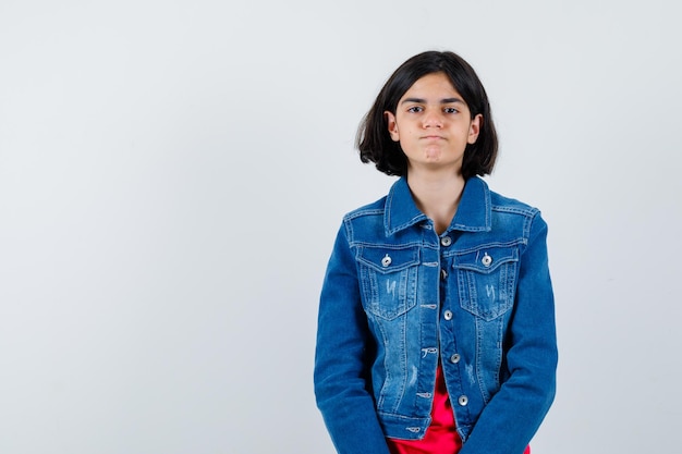 Young girl in red t-shirt and jean jacket standing straight and posing at camera and looking cute