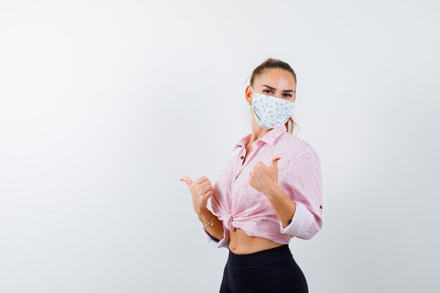 Young girl showing double thumbs up in pink blouse, black pants, mask and looking happy , front view.