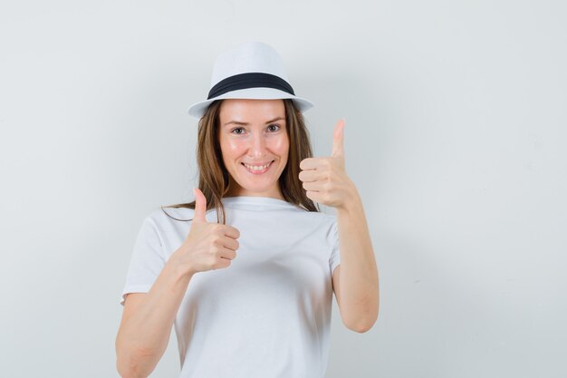 Young girl showing double thumbs up in white t-shirt, hat and looking glad , front view.