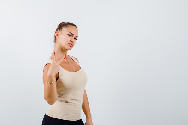 Young girl showing peace sign in beige top, black pants and looking confident. front view.