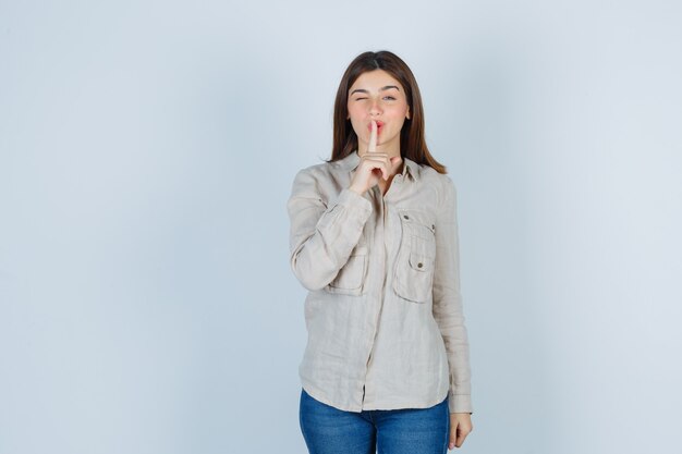 Young girl showing silence gesture, winking in beige shirt, jeans and looking cute. front view.