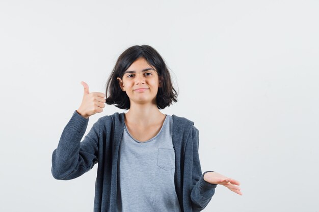 Young girl showing thumb up and stretching hand in light gray t-shirt and dark grey zip-front hoodie and looking cute