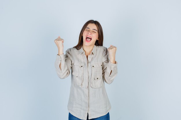 Young girl showing winner gesture in beige shirt, jeans and looking lucky , front view.