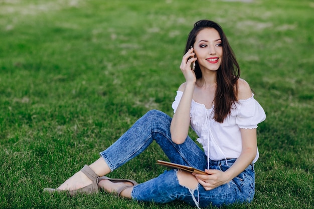 Young girl sitting in a park talking on a mobile while holding a tablet