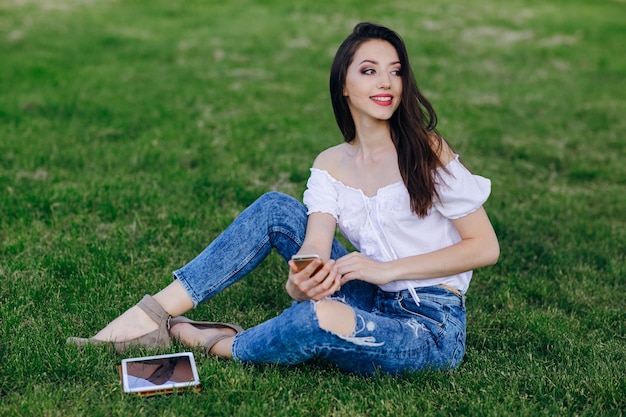 Young girl sitting in a park with a tablet in the grass and a mobile in the hand