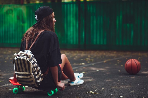 Young girl sitting on plastic orange penny shortboard on asphalt in cap