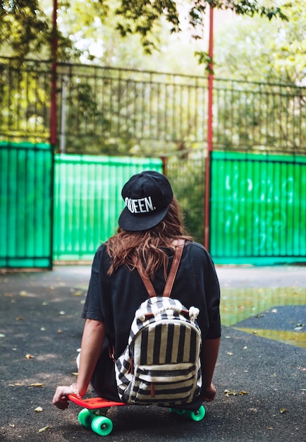 Young girl sitting on plastic orange penny shortboard on asphalt in cap