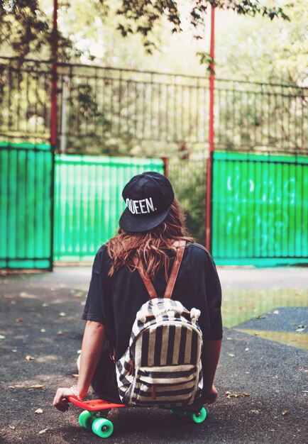 Young girl sitting on plastic orange penny shortboard on asphalt in cap