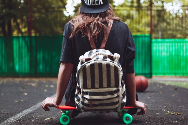 Young girl sitting on plastic orange penny shortboard on asphalt in cap