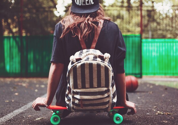 Young girl sitting on plastic orange penny shortboard on asphalt in cap