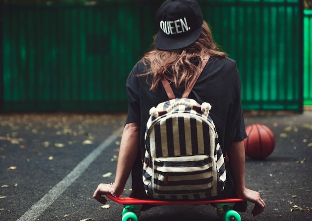 Young girl sitting on plastic orange penny shortboard on asphalt in cap