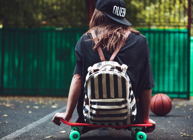 Young girl sitting on plastic orange penny shortboard on asphalt in cap