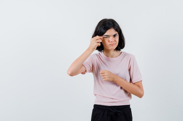 Young girl stretching eye with index finger in pink t-shirt and black pants and looking cute