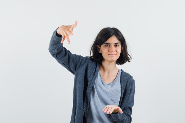 Young girl stretching hands as holding something big in light gray t-shirt and dark grey zip-front hoodie and looking cute.