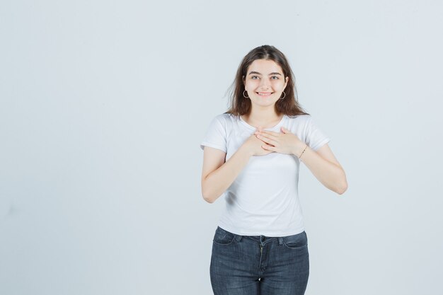 Young girl in t-shirt, jeans holding hands on chest and looking happy , front view.