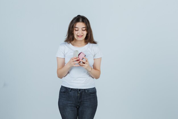 Young girl in t-shirt, jeans opening gift box while looking into it and looking amazed , front view.