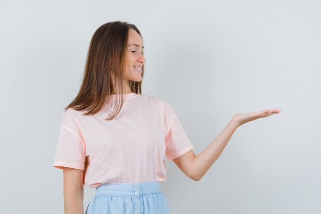 Young girl in t-shirt, skirt looking at palm spread aside and looking glad , front view.